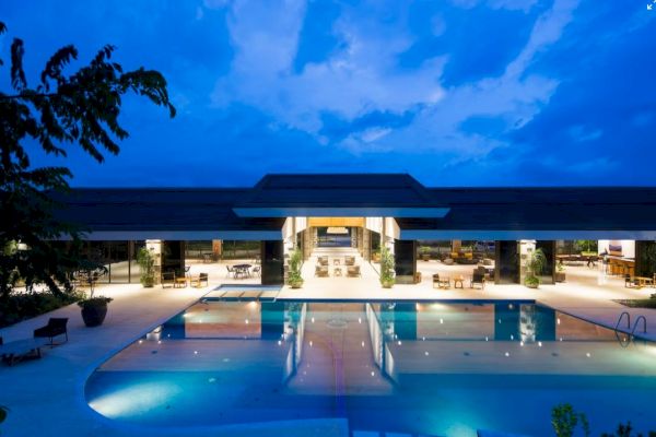 A luxurious building with a large, illuminated swimming pool at night, surrounded by lounge chairs and tables, under a partly cloudy sky.