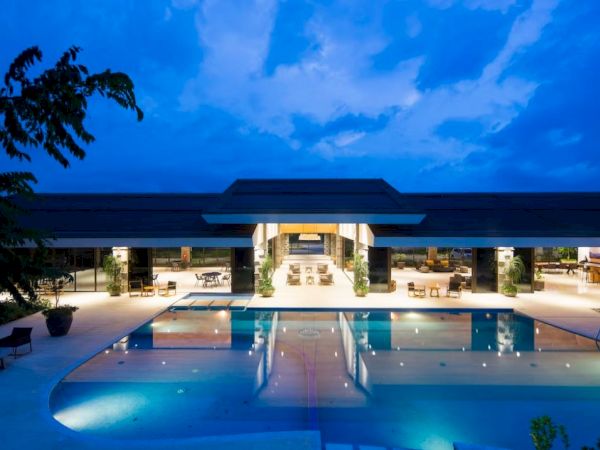 A luxurious building with a large, illuminated swimming pool at night, surrounded by lounge chairs and tables, under a partly cloudy sky.