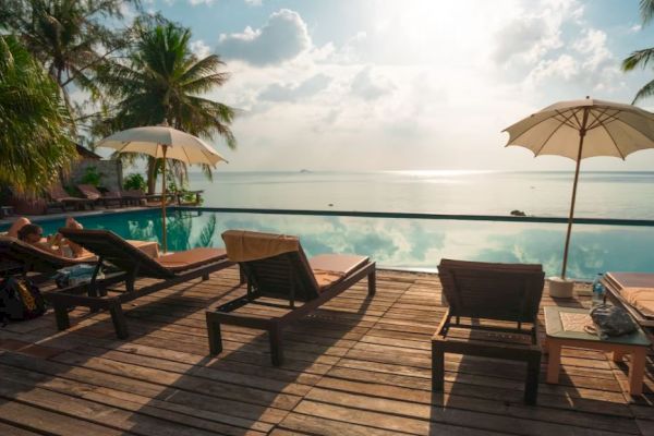A serene poolside scene with lounge chairs, umbrellas, and a tranquil view of the ocean and sky under the sunlight, surrounded by palm trees.