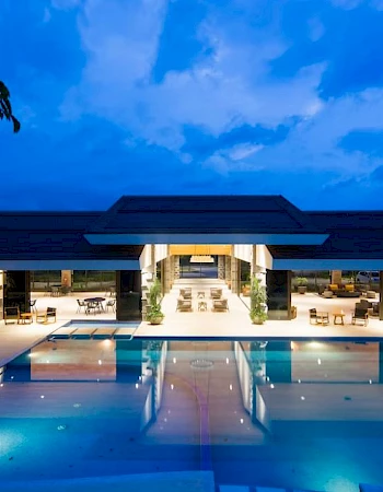 A luxurious, modern building with a large illuminated pool in front, surrounded by lounge chairs and greenery, under a vibrant evening sky.