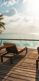 Poolside scene with lounge chairs, umbrellas, and a tranquil infinity pool overlooking the ocean, surrounded by lush palm trees and clear skies.