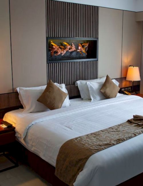 The image shows a hotel room with a large bed, two bedside tables with lamps, pillows, a folded blanket, and a wall-mounted decoration.