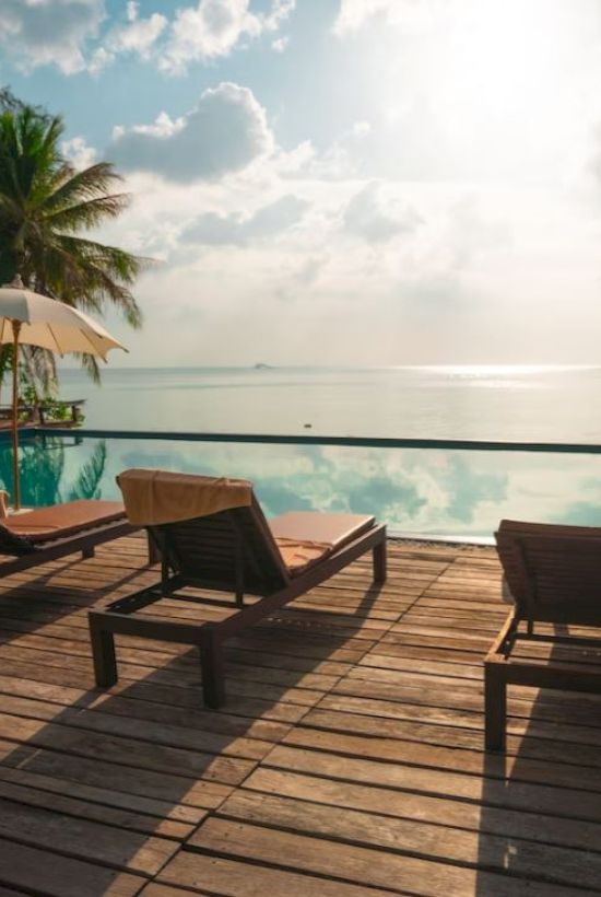 Lounge chairs and umbrellas by an infinity pool overlooking a serene ocean with palm trees in the background under a partly cloudy sky.