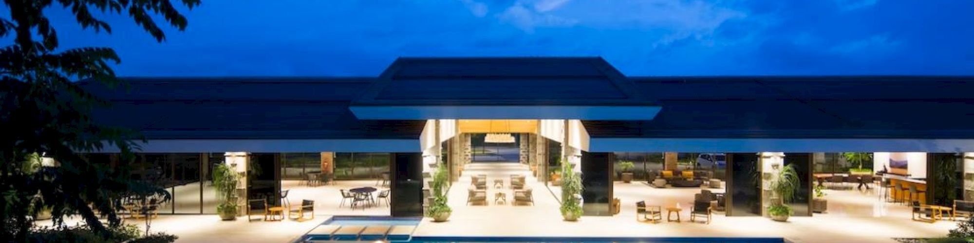 A luxurious resort featuring a large, illuminated swimming pool in front of an open, elegant building under a blue evening sky.