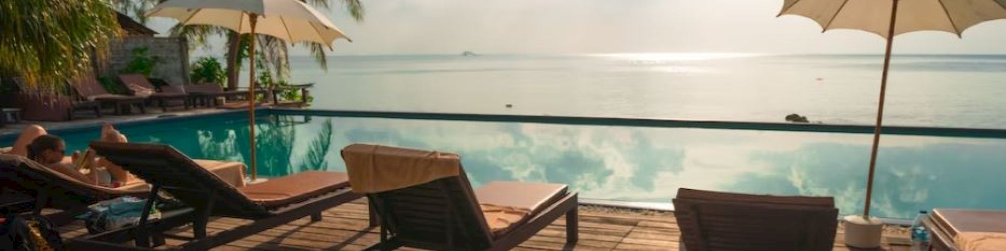 The image shows a serene poolside area with lounge chairs, umbrellas, and a view of a calm sea under a partly cloudy sky.