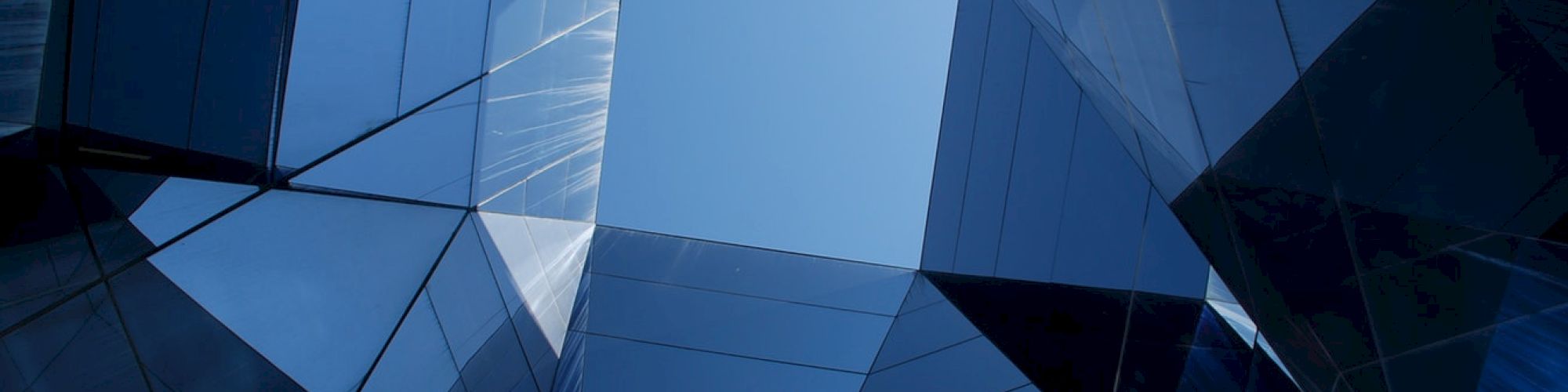 The image shows an abstract view looking upwards at a geometric, glass-walled structure, creating a pattern with reflections and the sky in the center.