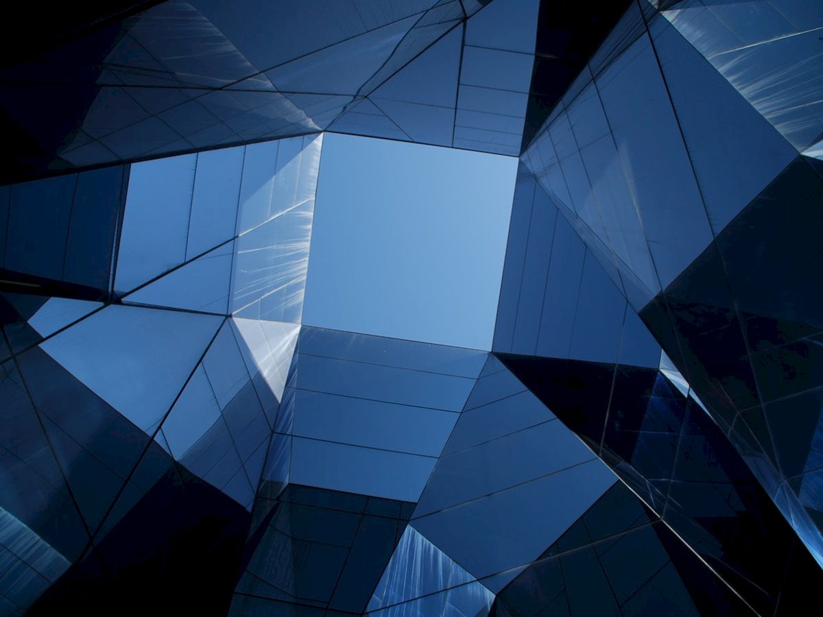 The image shows a geometric pattern of reflective glass panels forming a tunnel-like structure with a view of the sky at the top center.