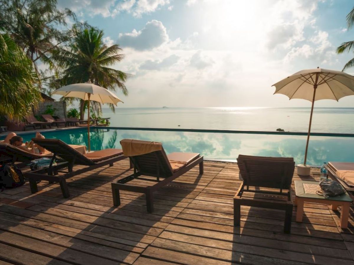A serene poolside scene with lounge chairs, umbrellas, and palm trees overlooking a calm, reflective sea under a bright sky.