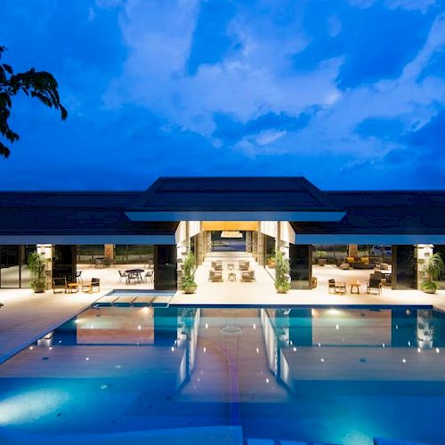 A well-lit modern building with a large pool in front, captured during dusk under a clear blue sky.