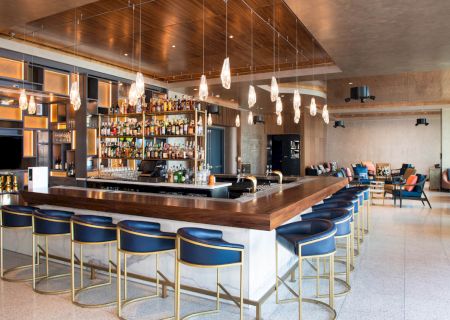 The image features a modern bar with a wooden countertop, stylish bar stools, hanging lights, and a well-stocked liquor shelf, enhanced by a cozy seating area.