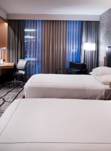 The image shows a modern hotel room with two neatly made beds, a flat-screen TV, a desk and chair, and contemporary lighting and decor.