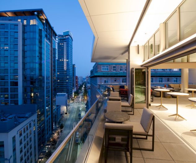 This image shows a modern city view at dusk from a high-rise building's outdoor terrace, featuring seating areas and a partially enclosed space.