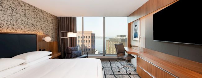 A modern hotel room with a large bed, flat screen TV, desk, and chair, offering a city skyline view through a large window.