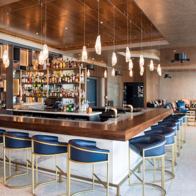 A modern bar with blue barstools, a wooden counter, hanging light fixtures, and a variety of bottles on shelves in a spacious, stylish setting.