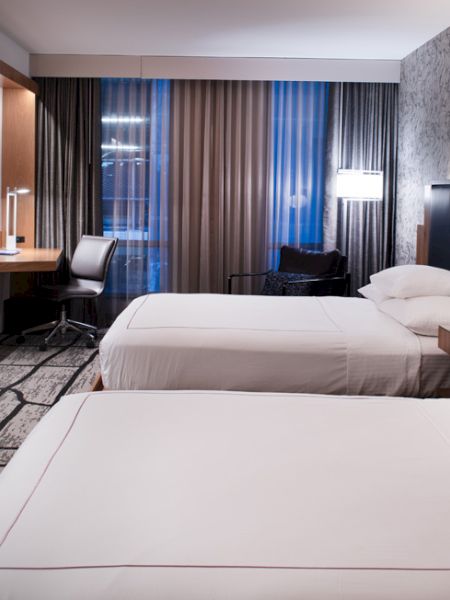 The image shows a modern hotel room with two neatly made beds, a flat-screen TV, a desk with a chair, floor-to-ceiling curtains, and wall-mounted lights.