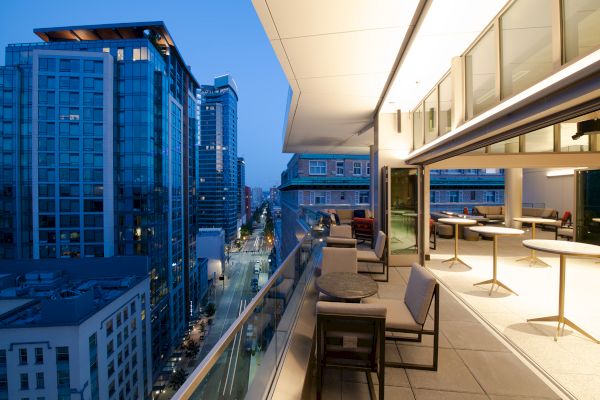 A modern cityscape at dusk with tall buildings, and a spacious balcony featuring chairs and tables overlooking the street below.