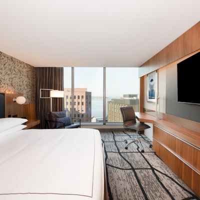 A modern hotel room features a king-sized bed, a large wall-mounted TV, a desk with a chair, and a window view of city buildings and water.