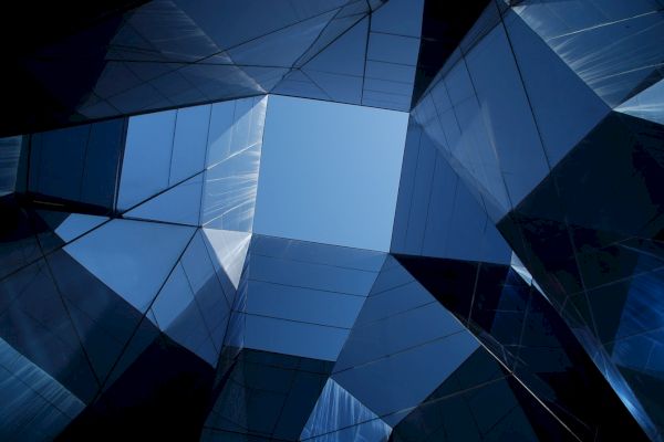 The image depicts a geometric arrangement of reflective glass surfaces forming an abstract structure with a patch of clear blue sky in the center.