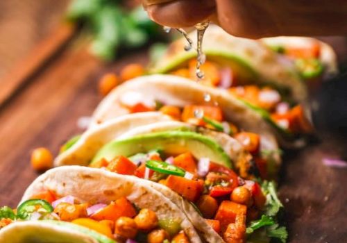 A hand squeezes lime juice over several vibrant vegetable tacos garnished with chickpeas, avocado slices, and cilantro on a wooden board.