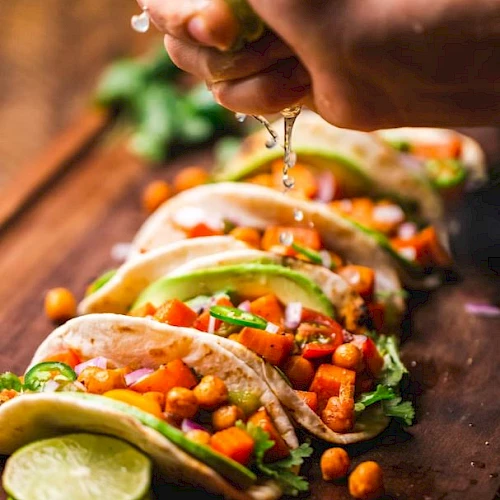 A hand squeezes lime juice over several vibrant vegetable tacos garnished with chickpeas, avocado slices, and cilantro on a wooden board.