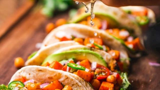 A hand squeezes lime juice over several vibrant vegetable tacos garnished with chickpeas, avocado slices, and cilantro on a wooden board.