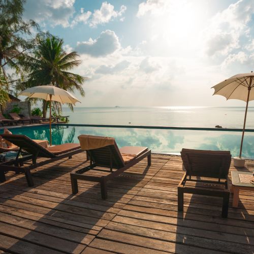 The image shows lounge chairs and umbrellas on a wooden deck by a serene pool facing the ocean with palm trees and a bright, sunny sky in the background.