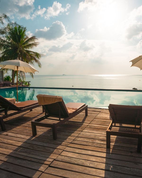 The image shows lounge chairs and umbrellas on a wooden deck by a serene pool facing the ocean with palm trees and a bright, sunny sky in the background.