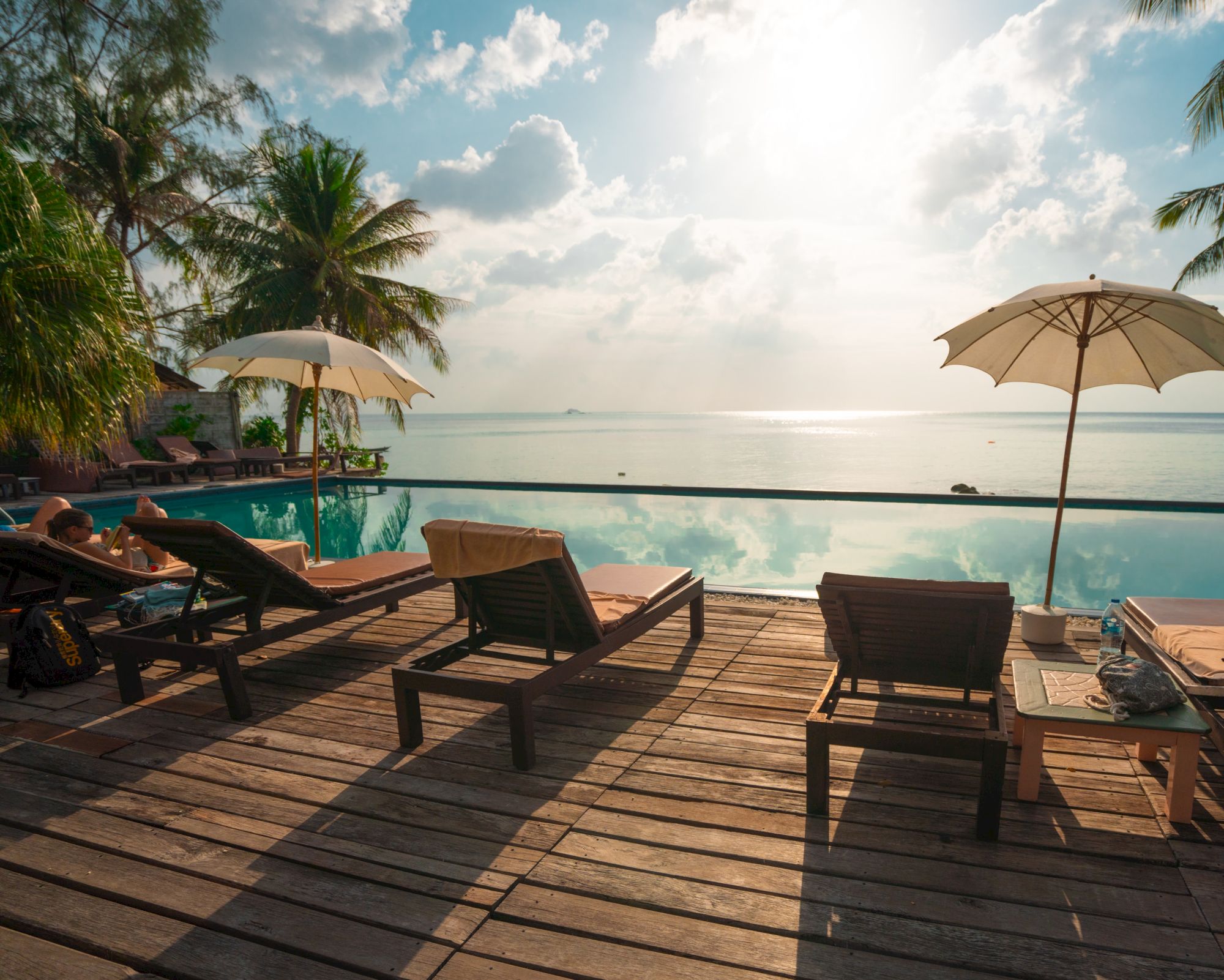 The image shows a serene beachside pool area with lounge chairs, umbrellas, and palm trees, overlooking a calm ocean under a sunny sky.