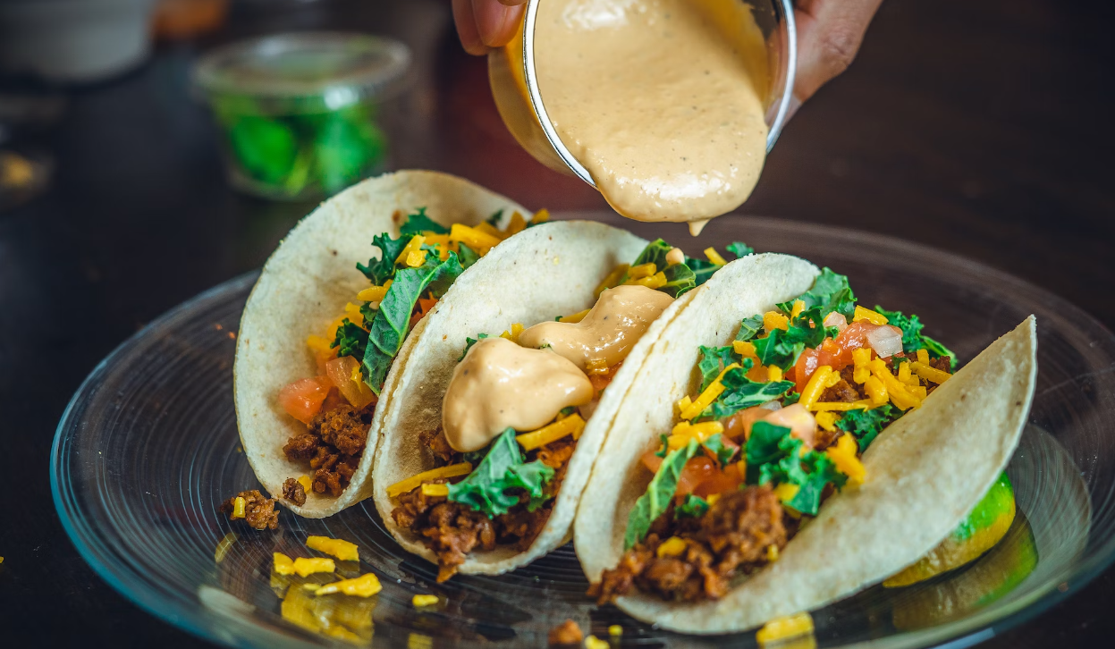 Three tacos filled with meat, lettuce, chopped tomatoes, cheese, and a hand pouring sauce atop on a clear plate.