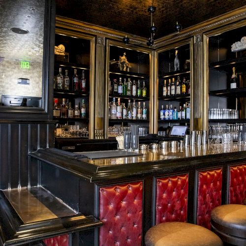 The image shows a stylish bar with red upholstered stools, polished wooden surfaces, and shelves stocked with various bottles and glassware.