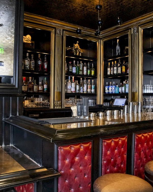 The image shows a stylish bar with red upholstered stools, polished wooden surfaces, and shelves stocked with various bottles and glassware.