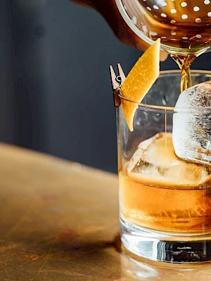 A hand is pouring a whiskey cocktail into a glass with ice and a lemon peel garnish on a bar counter.