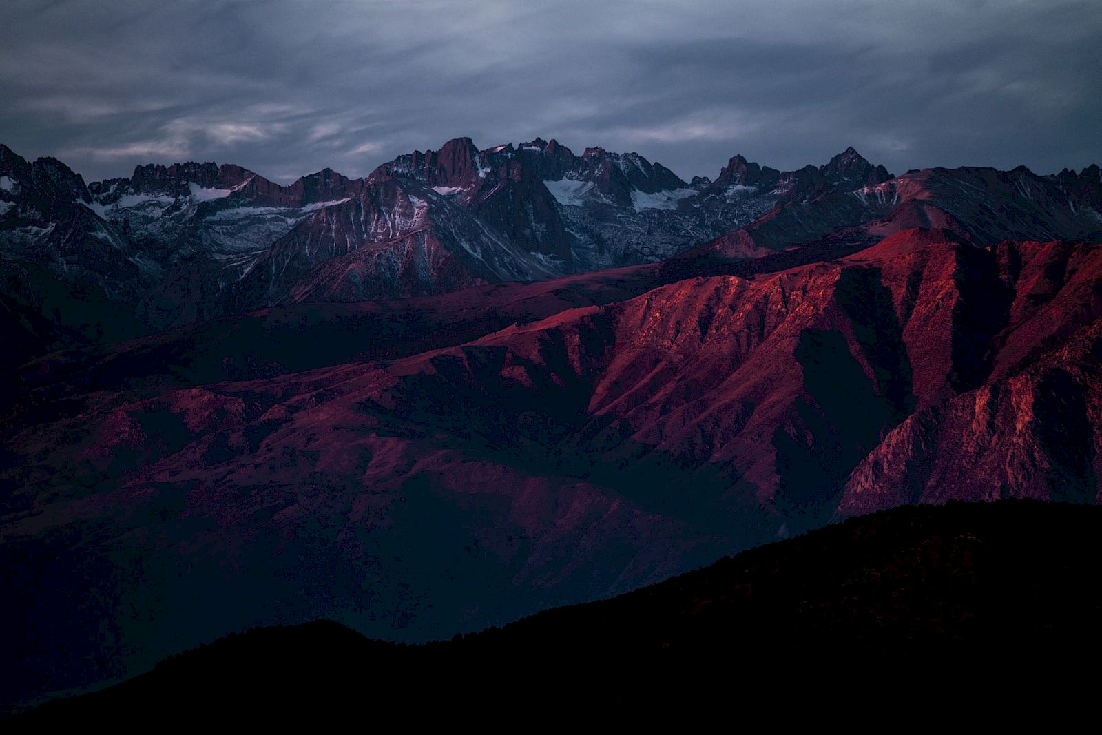 A mountain range is bathed in red hues under an evening sky, with snow-capped peaks in the background and dramatic lighting creating deep contrasts.