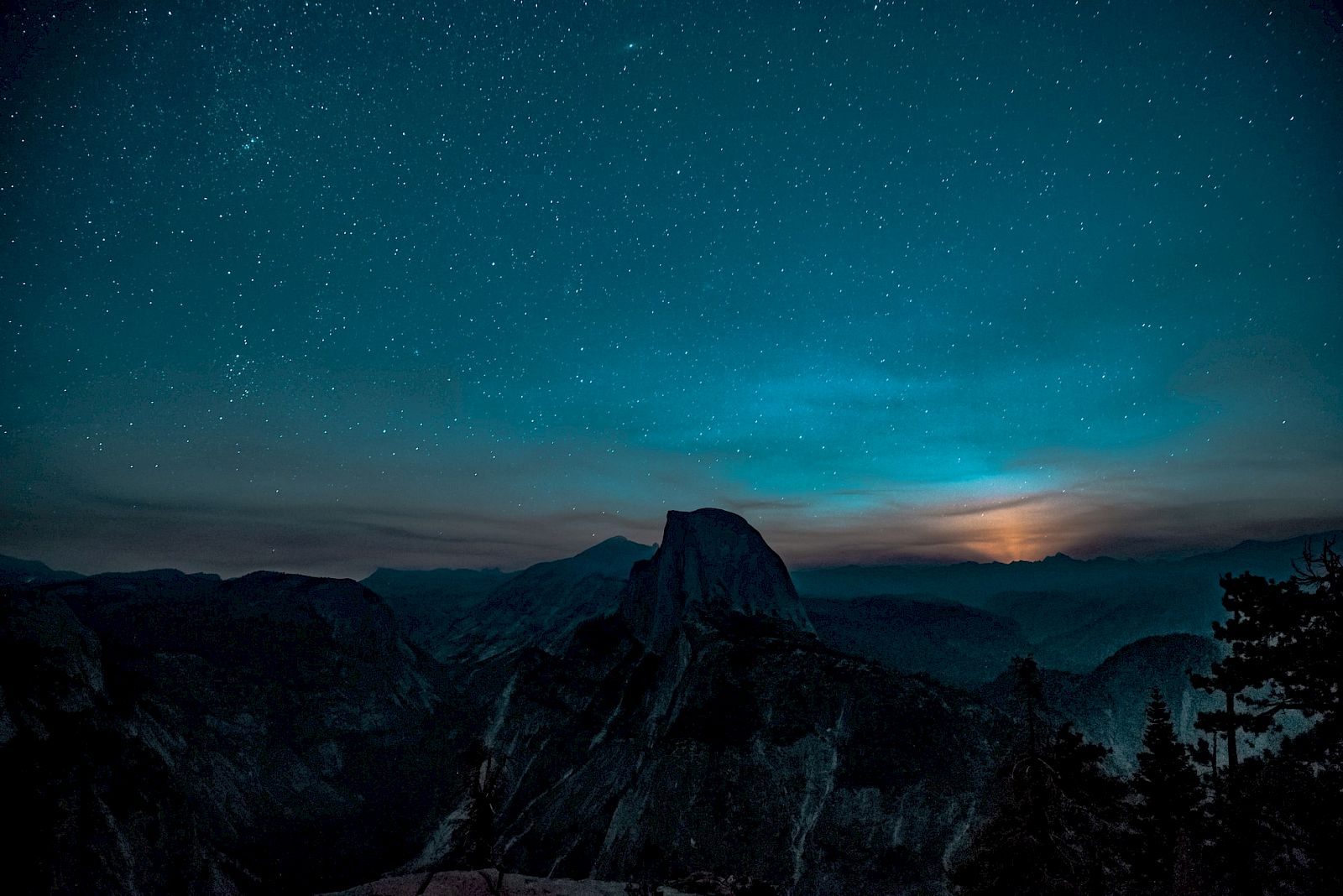 A stunning night sky filled with stars over a mountainous landscape, with a glowing light near the horizon and silhouettes of trees.