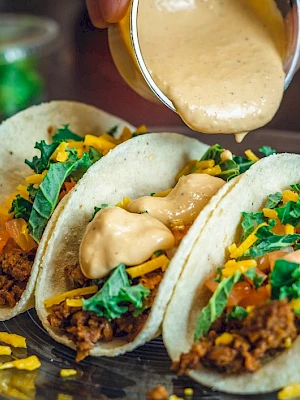 The image shows three tacos filled with ground meat, greens, cheese, and topped with a creamy sauce being poured from a small container.