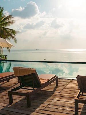 A serene beach scene with lounge chairs, umbrellas, palm trees, and a pool overlooking calm sea waters under a partly cloudy sky.
