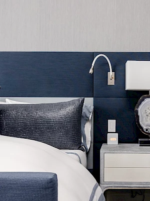 A modern bedroom featuring a neatly made bed with blue and white bedding, a nightstand with a lamp, and a wall-mounted reading light.