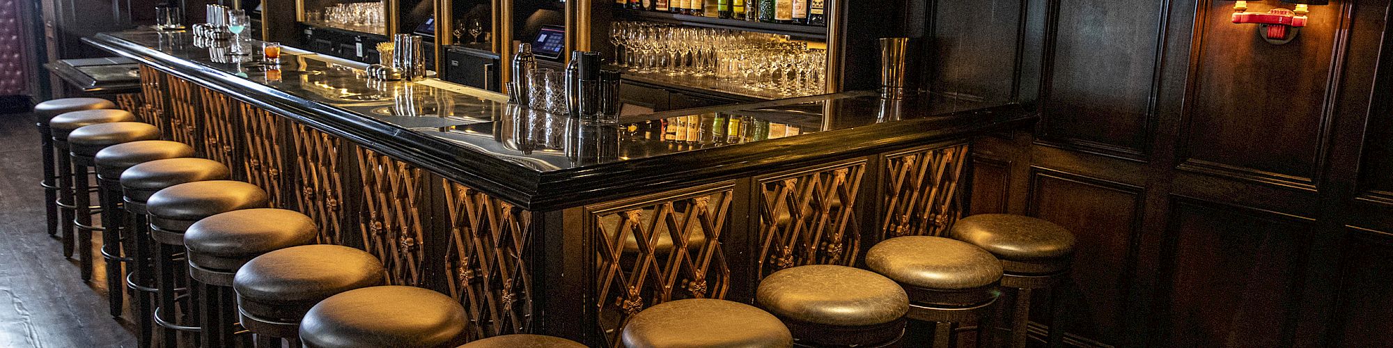 A dimly lit bar with a long counter, numerous bar stools, wooden interiors, and shelves stocked with various bottles of alcohol.