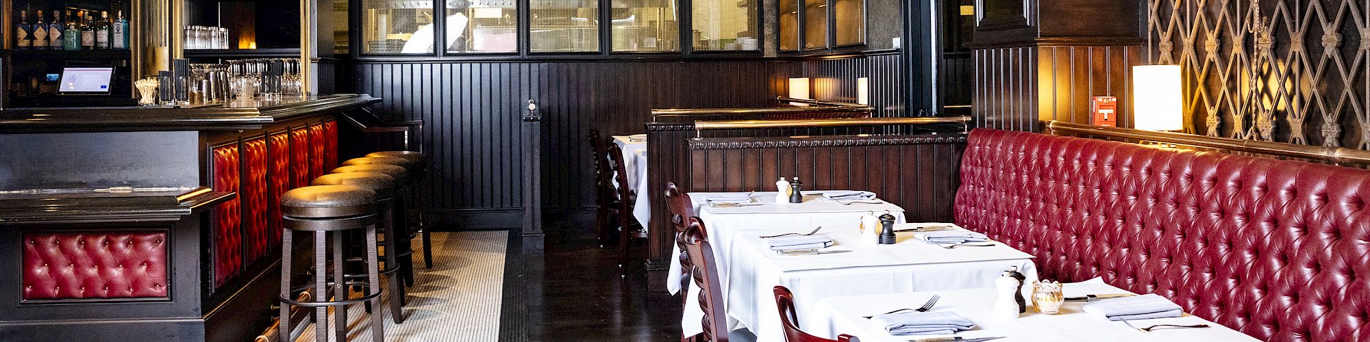 The image shows a restaurant interior with red tufted seats, white tablecloth-covered tables, a bar area with stools, and dim lighting.