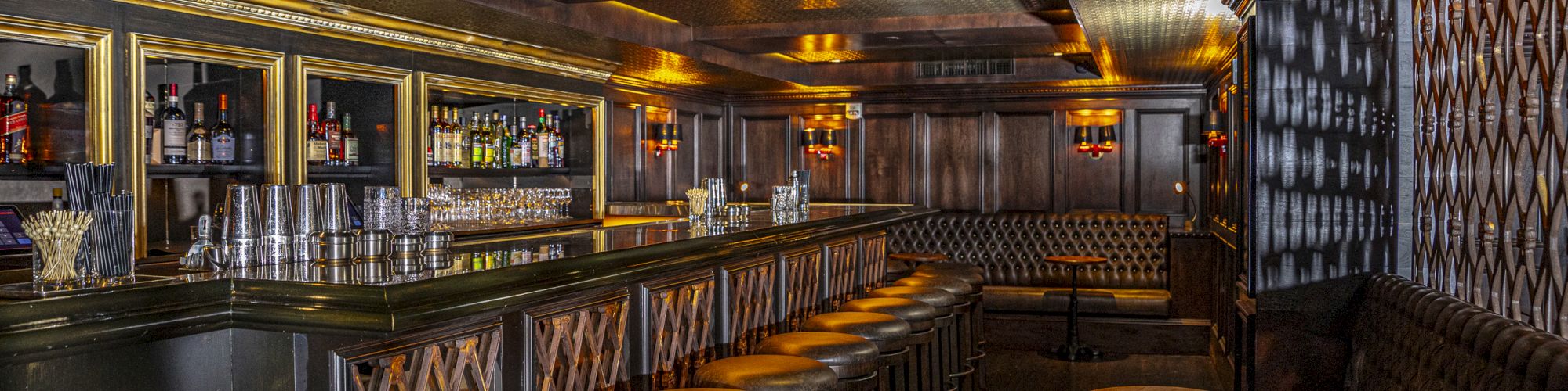 The image shows a dimly lit bar with stools, a counter, shelves with bottles, and round tables along the wall.