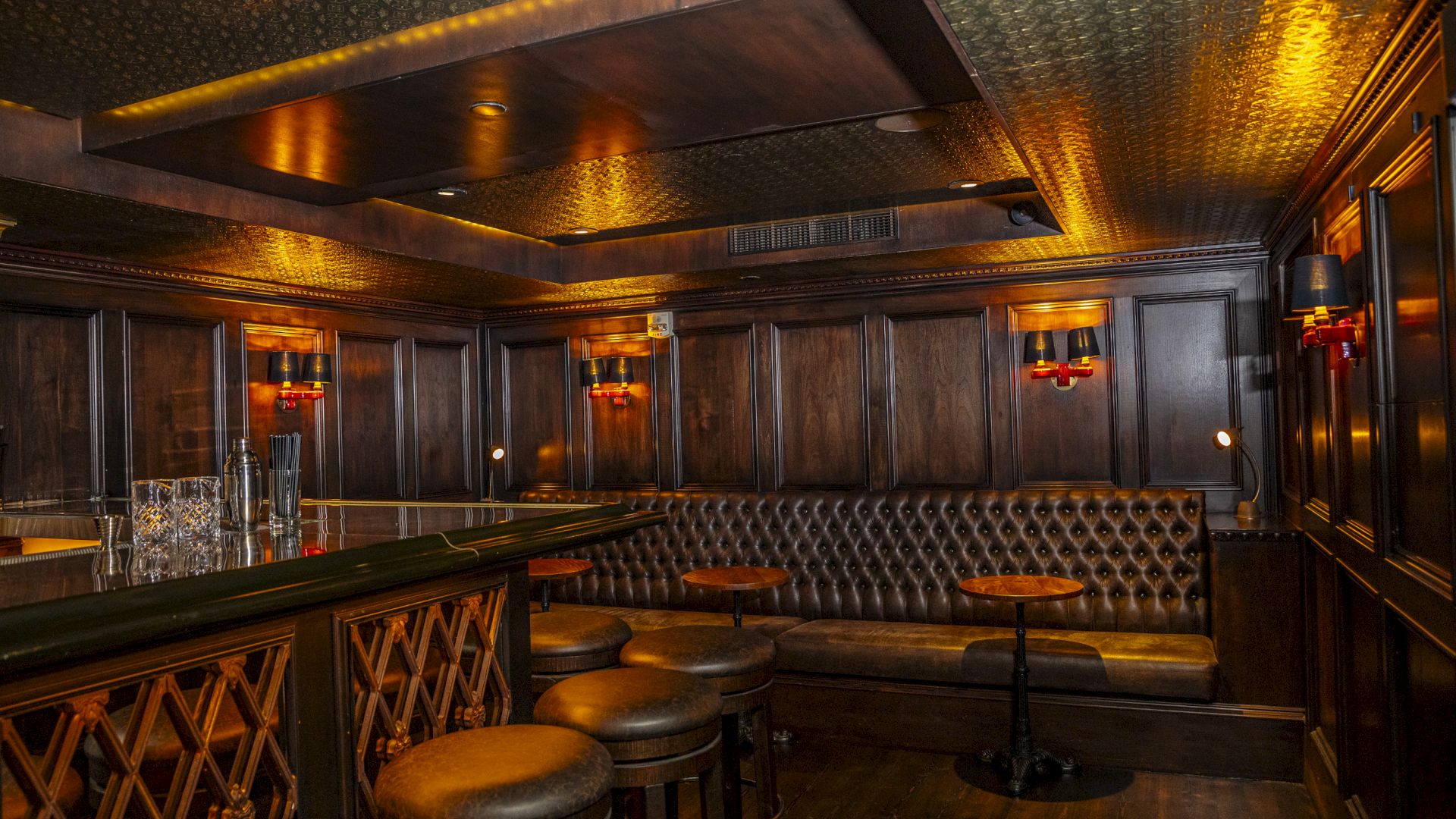 The image shows a dimly lit, cozy bar with leather seating, wooden accents, bar stools, and small round tables in an elegant, intimate setting.