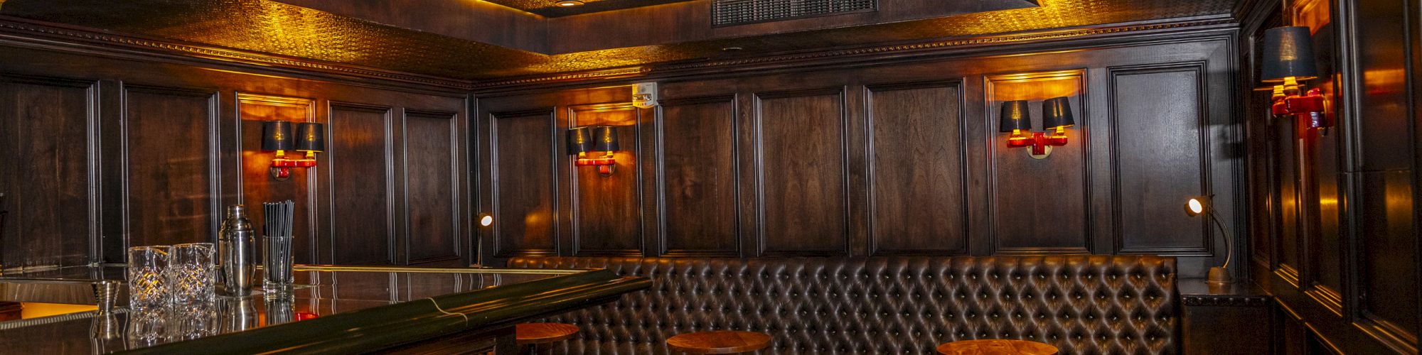 The image shows a dimly lit, cozy bar with leather seating, wooden accents, bar stools, and small round tables in an elegant, intimate setting.