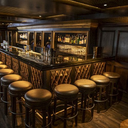 A dimly lit bar with a long counter, stocked shelves, and leather stools arranged neatly along the bar, creating an inviting atmosphere.