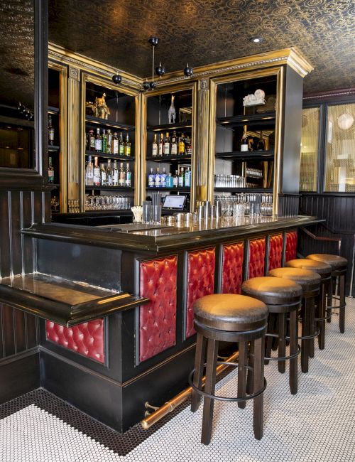 A stylish home bar with red upholstered panels, wooden bar stools, shelves stocked with bottles, and a polished countertop.