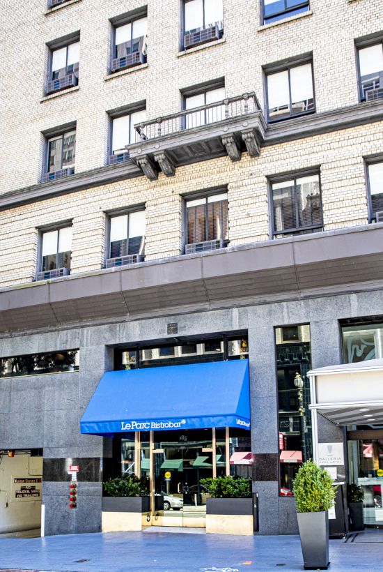 The image shows the exterior of a multi-story building with a blue awning above the entrance and plants flanking the door.