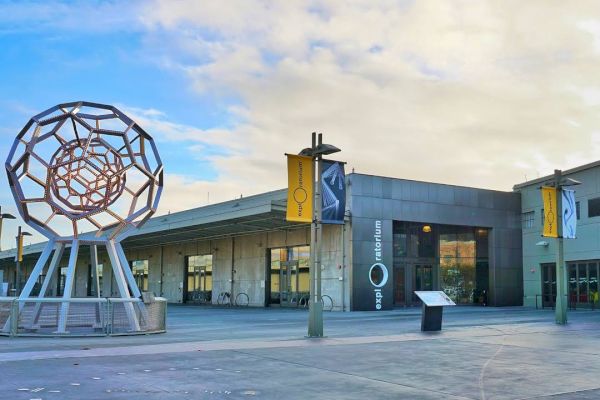 The image shows a modern building with geometric sculpture outside and yellow banners on poles. The building has signs that read 