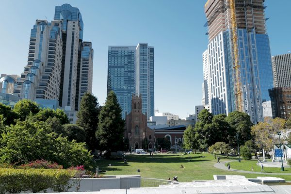 A cityscape featuring a mix of modern skyscrapers and a historic church, surrounding a green park area with people walking and relaxing.