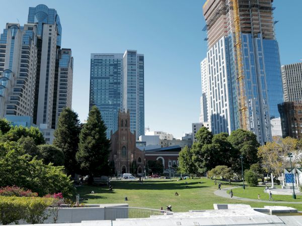 A cityscape featuring a mix of modern skyscrapers and a historic church, surrounding a green park area with people walking and relaxing.