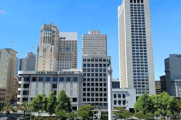 The image features a cityscape with tall buildings, a plaza with a monument at the center, and greenery. The sky is clear and blue.