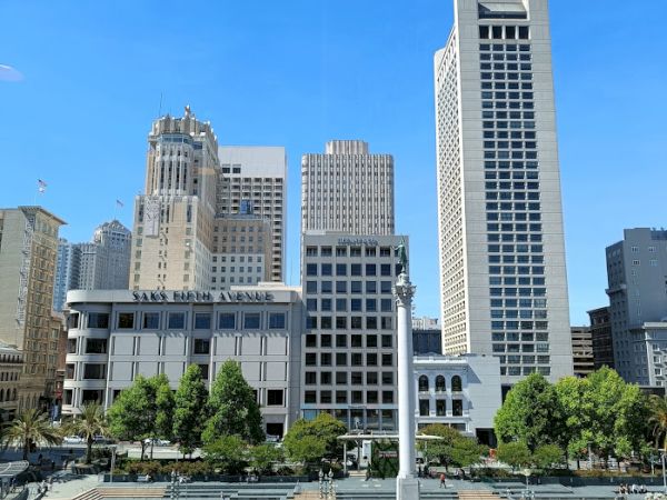 The image features a cityscape with tall buildings, a plaza with a monument at the center, and greenery. The sky is clear and blue.